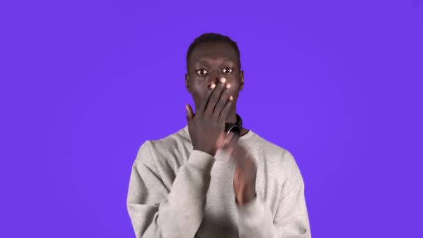 Glada ung afrikan man, gestikulerar mot någon. Isolerad på blå bakgrund. Bär vit tröja och hörlurar på halsen — Stockvideo