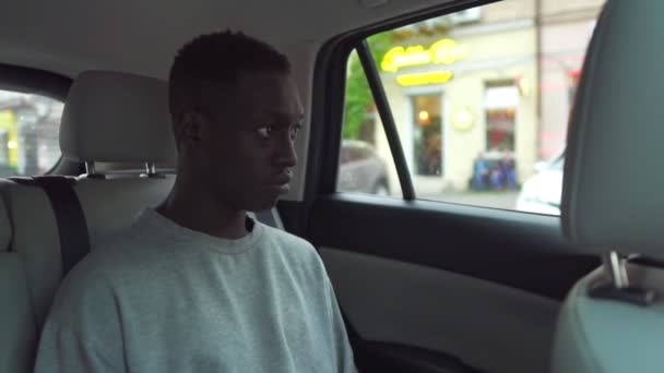 Guapo joven negro cabalga en el asiento trasero de un coche, mira a su alrededor maravillado por la ventana. Gran vista de la ciudad reflejada en ventana. Vestido con camisa blanca casual. Movimiento lento — Vídeo de stock