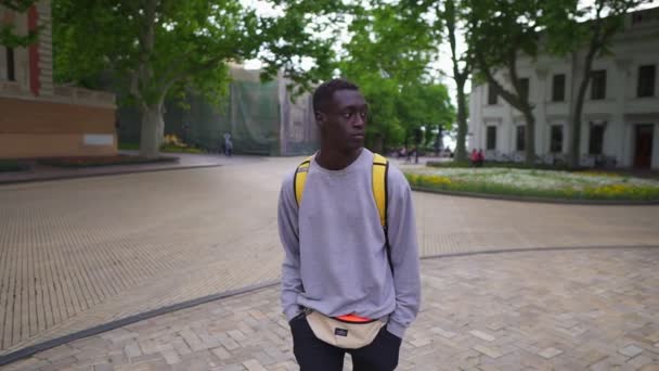 Jóképű, fiatal afro-amerikai férfi besétál a városba sárga hátizsákkal a vállán, alkalmi ruhát visel. Nézz körül, régi utcák háttere. Előnézet — Stock videók
