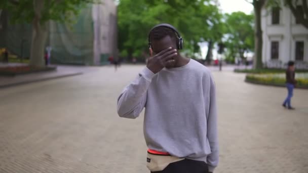 Langzame beweging van vrolijke Afro-Amerikaanse man in casual kleding dansen, vrij bewegend in hoofdtelefoon buiten plezier in de stad alleen, glimlachend, zorgeloos. Jongeren- en amusementsconcept — Stockvideo