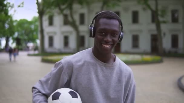 Afroamerikanischer Fußballspieler steht mit einem Fußballball in der Hand auf einer Straße in der Altstadt. Positiver Typ mit Kopfhörern und lässiger Kleidung, der in die Kamera lächelt — Stockvideo