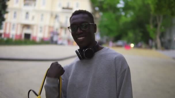 Retrato de jovem afro-americano em óculos elegantes e fones de ouvido olhando para o sorriso da câmera se sentir feliz. Coloque em sua mochila, cara de pessoas de moda felicidade, bonito atraente câmera lenta urbana — Vídeo de Stock