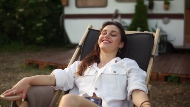 Agréable, jolie femme caucasienne se rafraîchissant et souriant tout en s'allongeant sur une chaise longue à l'extérieur. Voyage, repos, concept de vacances. Remorque, roulotte maison sur le fond flou — Video