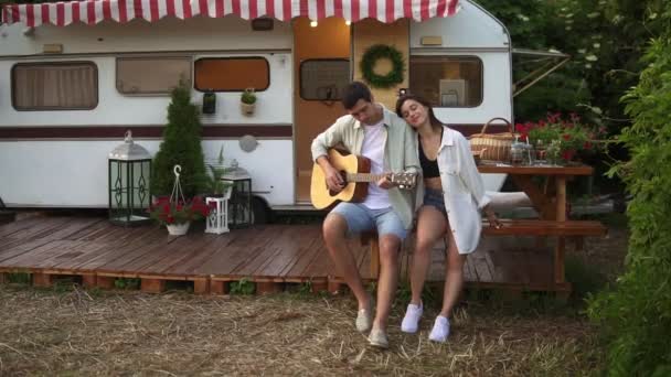Příjemný, uvolněný pár sedící venku před svým domácím přívěsem a muž hrající na kytaru pro přítelkyni. Venkovní. Dívka poslouchající hudbu a objímající muže. Zpomal. Portrét — Stock video