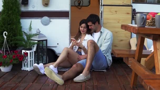 魅力的なカップルは、ドアのバンの近くの床に座って、スマートフォンでビデオを見て、笑顔。一緒に楽しむ、休日を共有し、車輪の家で旅行する — ストック動画