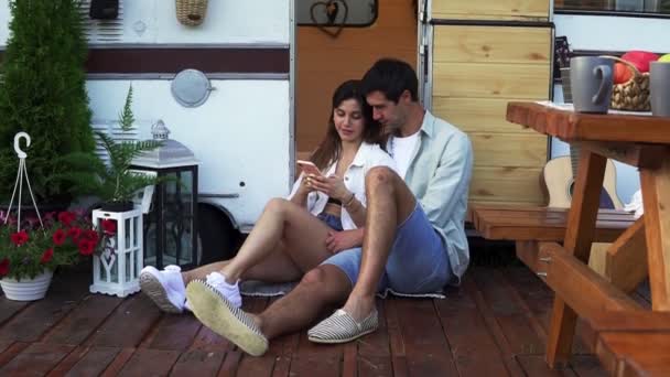 Atractiva pareja sentada en el suelo cerca de la furgoneta de la puerta y viendo el vídeo en el teléfono inteligente, sonriendo. Disfrutar juntos, vacaciones compartidas, viajar en casa de ruedas — Vídeo de stock