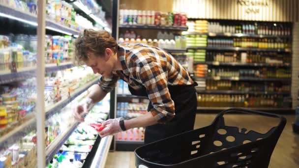 Verkäufer stellt Käsepackungen aus dem Einkaufswagen in die Regale der Milchprodukteabteilung im Lebensmittelladen, ein bärtiger Mann trägt Schürze. Verkauf von Produkten, Beruf und Handelskonzept — Stockvideo