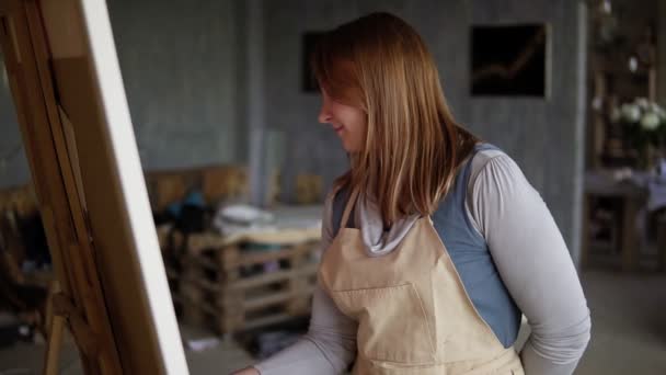 Retrato de una mujer caucásica pintando en un estudio de artes usando manchas y pinturas al óleo. Artista de pelo largo en delantal beige, sonriendo durante el proceso, mirando a la cámara. Movimiento lento — Vídeo de stock