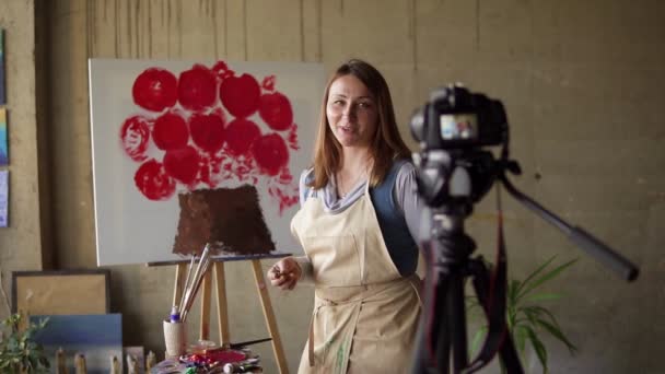 Mujer caucásica madura blogger grabación de vídeo en el taller - dibujo de flores de color rosa sobre lienzo, explicando los detalles en la cámara. Movimiento lento — Vídeos de Stock