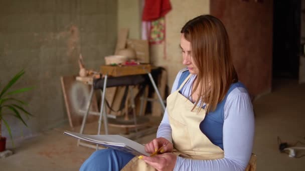 Artista mujer en el lugar de trabajo dibujo bosquejo lápiz. Mujer de pelo largo sentada en la silla en el lugar de trabajo, sosteniendo la hoja de trabajo en las rodillas — Vídeo de stock