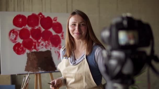 Female caucasian mature blogger recording video in workshop - drawing pink flower on canvas, explaining details on camera. Slow motion. Tripod camera on foreground — Stock Video