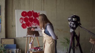 Beyaz kadın yetişkin blog yazarı atölyeye video kaydediyor - tuvale pembe çiçek çiziyor, kameradaki detayları anlatıyor