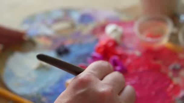 Die Künstlerin mit dem dicken Pinsel mischt verschiedenfarbige Ölfarben auf Palette - lila, rot, gelb. Nahaufnahme weiblicher Hand beim Mischen von Farben. Bunte Palette — Stockvideo