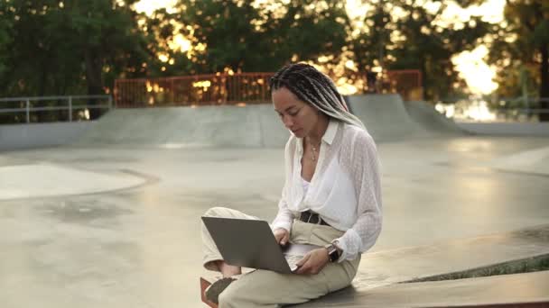 Joven mujer caucásica elegante con rastas en blusa blanca sentado en el parque de verano. Feliz freelancer escribiendo en el portátil al aire libre en skatepark — Vídeos de Stock