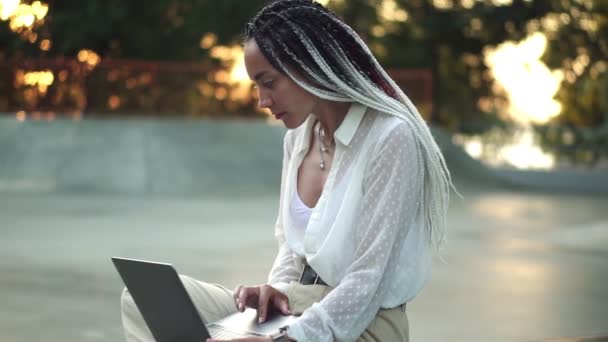 Vista laterale di bella, ragazza elegante con dreadlocks in bianco e nero seduto sul parapetto nello skate park locale che lavora sul computer portatile, digitando. Indossando bella camicetta bianca e pantaloni beige, Bike rider on — Video Stock