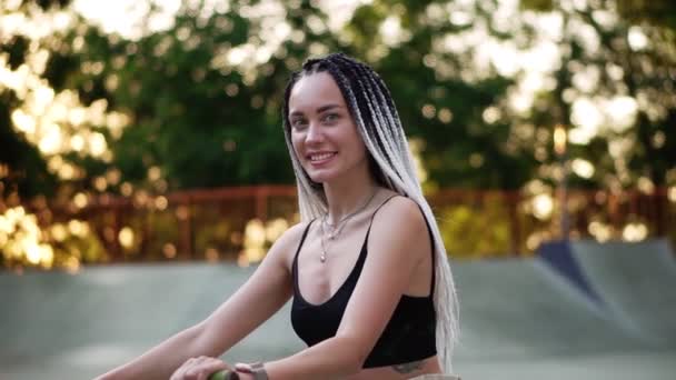 Menina com dreadlocks preto e branco sentado na moto no parque de skate e sorrindo para a câmera. Árvores borradas à luz do sol no fundo — Vídeo de Stock