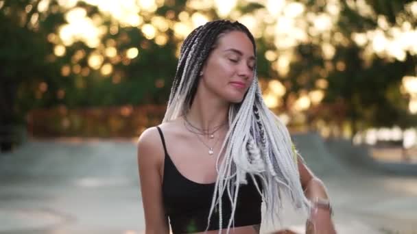 Mulher sensual em top preto com dreadlocks preto e branco dançando no skatepark vazio. Menina dançando relaxantemente com os olhos fechados. A usar decote prateado. Árvores à luz do sol no fundo — Vídeo de Stock