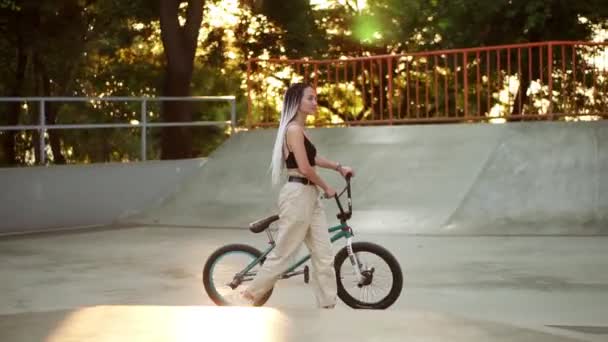 Muchacha atractiva con estilo con rastas paseos con bicicleta bmx en skatepark en un parque de recreo. Mujer joven y elegante en bicicleta en la ciudad. Parque de verano vacío. Longitud completa, vista lateral — Vídeo de stock