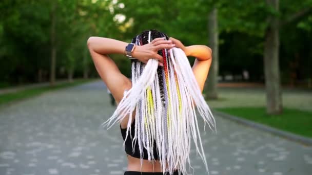 Schwarz-weiße Dreadlocks auf dem Rücken eines Mädchens. Rückansicht eines sportlichen Mädchens, das Haare knüpft und im grünen, öffentlichen Park zu laufen beginnt — Stockvideo
