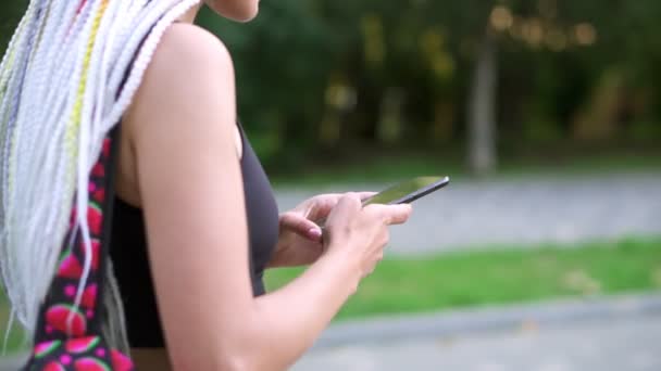 Gadis tak dikenal dengan pesan ransel menggunakan smartphone. Wanita dengan rambut gimbal berjalan di jalan, taman hijau dalam gerakan lambat. Tampilan samping — Stok Video