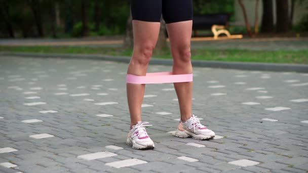 Giovane atleta irriconoscibile esercita con banda di resistenza rosa facendo squat all'aperto al parco pubblico locale, verde. Donna attraente che indossa abbigliamento sportivo nero e sneaker. In piedi sul marciapiede — Video Stock