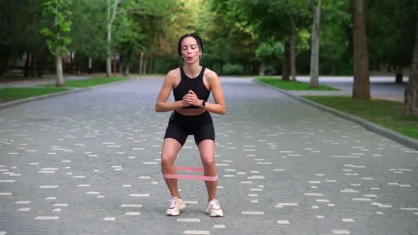 Front view of stylish woman with dreadlocks makes crounchy side steps with pink rubber band on legs on pavement, view on green city park on background. Breathing correctly — Stock Video