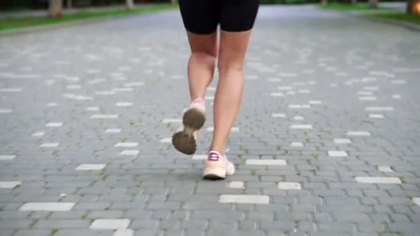 Visão traseira da mulher europeia com dreadlocks preto e branco executando por local, parque verde na cidade. Ela está se exercitando para uma boa saúde. Vestindo top preto e shorts. Fechar as pernas dos atletas — Vídeo de Stock