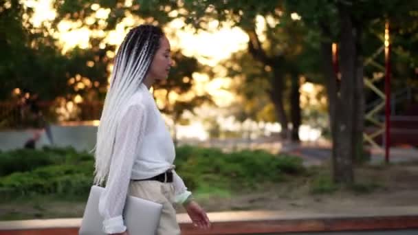 Zelfverzekerde wandeling van een elegant meisje in witte blouse en broek - jong meisje loopt door het park met een laptop in haar hand. Zijaanzicht. Lenzen fakkels — Stockvideo