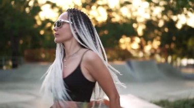 Parkta dans eden Afrikalı saçlı güzel bir kız. Güneşli bir günde siyah bluzlu ve güneş gözlüklü güzel bir kadın rahatça dans ediyor. Yavaş çekim yap. Bulanık arkaplan
