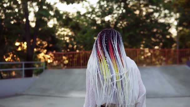 Carefree woman in casual with colorful dreadlocks standing in the center of outdoors skatepark. Girl in light white blouse dancing and turning around against sun with trees on background. Slow motion — Stock Video