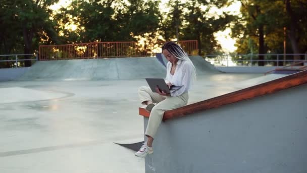 Fiatal, elegáns fehér nő, fehér blúzban, aki a nyári parkban dolgozik. Női szabadúszó gépelés laptop szabadban ül masszív korlátok skatepark — Stock videók