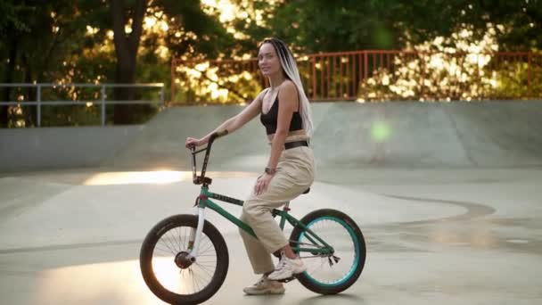 Portret pięknej dziewczyny z czarno-białymi dredami siedzącej na rowerze w skateparku i uśmiechniętej do kamery. Zamazane drzewa w złotym słońcu na tle — Wideo stockowe