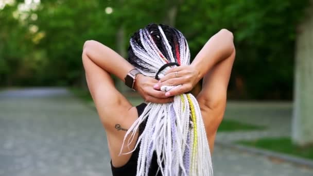 Zwart-witte dreadlocks op de rug van een meisje. Achteraanzicht van een sportief meisje kammen haar met haar rubber voor comfortabele wandeling of run. Zomer, groen park — Stockvideo