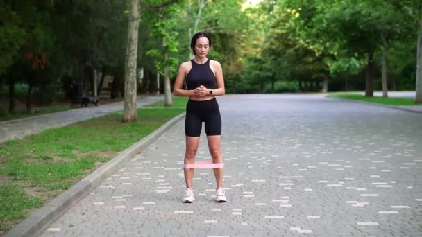Front view of stylish woman makes side steps with pink rubber band on legs on pavement, view on green city park on background, slow motion. Outdoor fitness workout — Stock Video