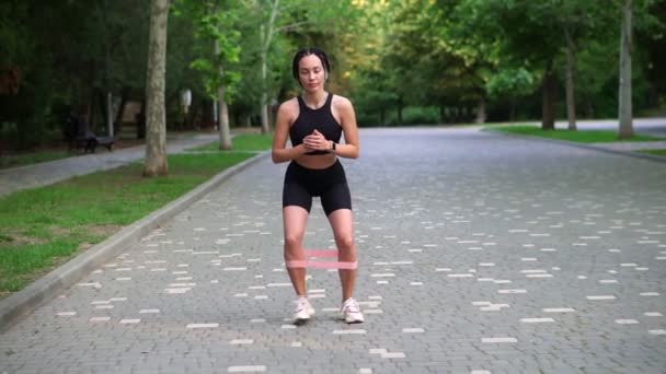 Front view of stylish woman makes crounchy side steps with pink rubber band on legs on pavement, view on green city park on background, slow motion. Outdoor fitness workout — Stock Video