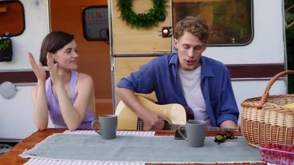 Positive couple sitting at the picnic table outdoors in front their home trailer and man playing the guitar and sings song actively. Girl listening music and clapping hands. Slow motion — Stock Video