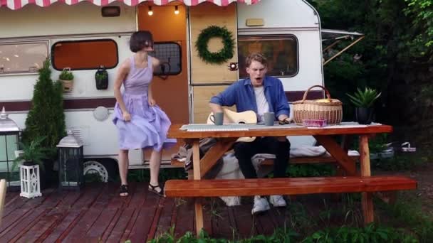 Menina morena em azul vestido de verão menina dançando enquanto o homem tocando guitarra sentado na mesa de madeira no trailer da frente, cantando uma música em voz alta. Férias, férias, viagem de caravana. Movimento lento — Vídeo de Stock