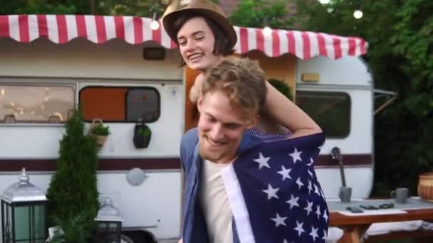 Despreocupado, alegre pareja divirtiéndose cerca de remolque en el parque, hombre piggybacking chica con bandera americana en su espalda girándola alrededor — Vídeo de stock