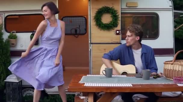 Смішна дівчина в блакитній літній сукні танцює, поки чоловік грає на гітарі, сидячи за дерев'яним столом перед трейлером, голосно співаючи пісню. Відпустка, відпустка, поїздка на трейлер — стокове відео