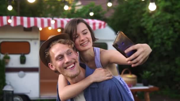 Parktaki karavanın yanında eğlenen mutlu genç çift, sırtına binen adam. Yazlık elbiseli ve şapkalı gülümseyen kız akıllı telefon kullanarak selfie çekiyor. Neşeli, genç bir çiftin mükemmel fotoğrafı. — Stok video