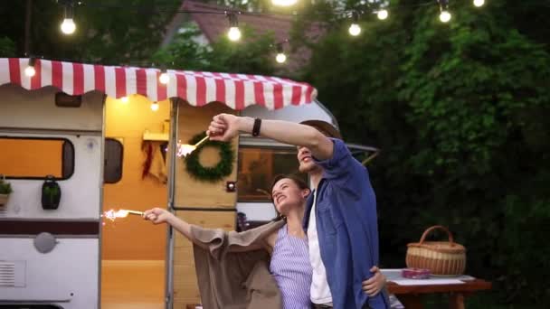 Young couple embracing standing in front trailer they living in with sparklers celebrating, waving hands with fireworks sticks. Happy People. Slow motion — Stock Video