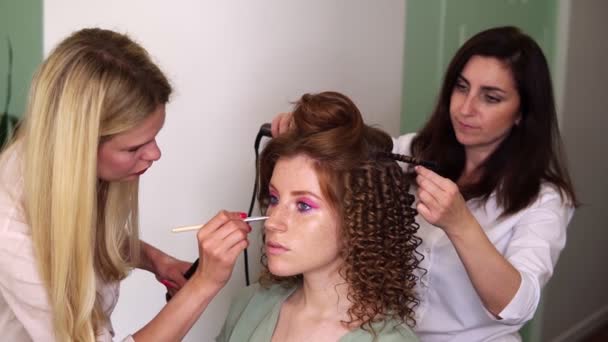 Två konstnärer arbetar med klienten i tandem - makeup artist tillämpar toner i ansiktet på en vacker modell med fräknar, röd huvud. Frisör med en locktång gör frisyr. Långsamma rörelser — Stockvideo