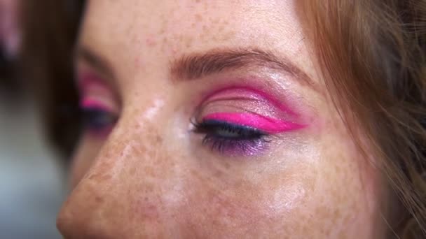 Nierozpoznawalny charakteryzator pracuje w studiu kosmetycznym. Kobieta stosująca ton pędzlem. Artysta zrobić makijaż dla rudej modelki z piegami. Zbliżenie oczu ładnej dziewczyny z różowym połyskiem makijażu — Wideo stockowe