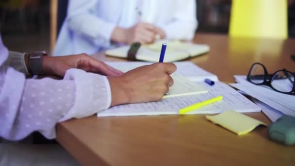 Una mujer escribe notas en el bloc de notas. Escribir ideas de negocios. Primer plano de dos mujeres de negocios escribir notas a mano. Mujeres irreconocibles en blusas blancas sentadas a la mesa de madera — Vídeos de Stock