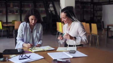 Masada oturan, el yazısı yazan, fikirleri paylaşan iki çekici, beyaz kadın. Beyaz bluzlu kadınlar modern kütüphanede oturuyor, proje üzerinde birlikte çalışıyorlar.