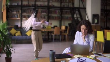 Halk kütüphanesini aç. Mutlu rastalar beyaz bluz ve bej pantolon giyen güzel kız arka planda dans ederken arkadaşı onun çalışmasına konsantre olmuş, masada dizüstü bilgisayarla çalışıyor.