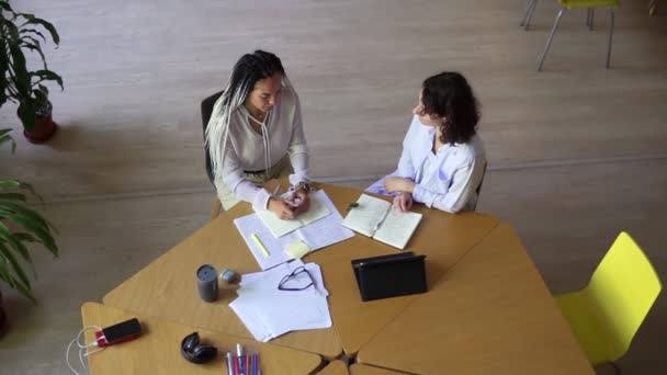 Duas mulheres conversando em uma biblioteca com prateleiras de livros em segundo plano. Sentado na mesa de madeira com tablet e papéis diversos sobre ele. Menina com dreads elegantes explicando smth. Topo — Vídeo de Stock