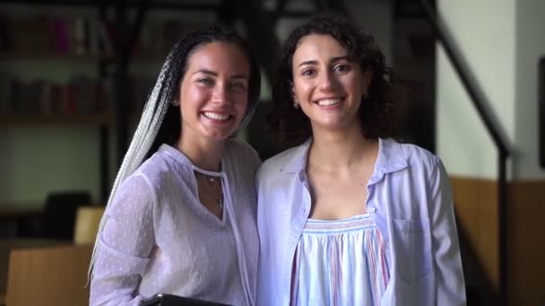 Портрет двох красивих білих жінок у білих блузах, що стоять у бібліотеці та посміхаються у камері, академічні друзі — стокове відео