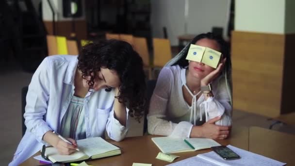 Duas mulheres caucasianas a gozar durante a preparação para os exames. Menina engraçada com dreadlocks sentado tendo papel amarelo adesivo com olhos pintados, enquanto sua amiga está escrevendo seu caderno — Vídeo de Stock