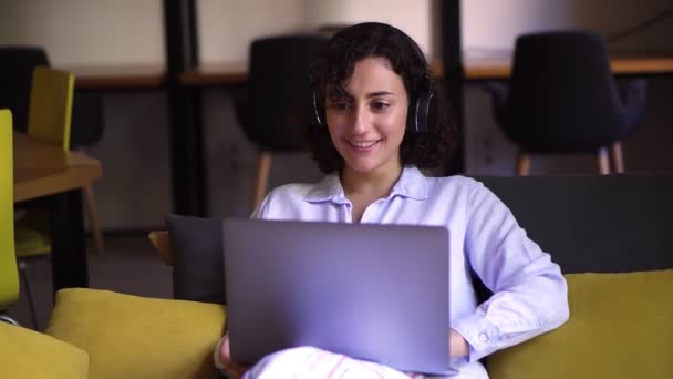 Portræt af krøllet, brunette kvinde, der arbejder på laptop på hjemmekontoret. Ung kvindelig professionel arbejder på computer enhed. Fjernbetjening freelance arbejde eller studere - i hovedtelefoner, smilende – Stock-video